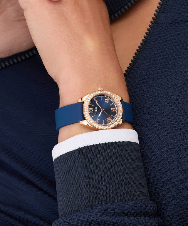 Wrist wearing a GUESS Ladies Mini Luna Blue Rose Gold Tone Analog Watch with a gold case on a dark background