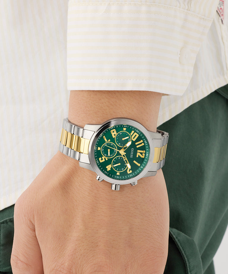 Green watch with silver and gold accents on a wrist, worn by a person in a white shirt and green pants.