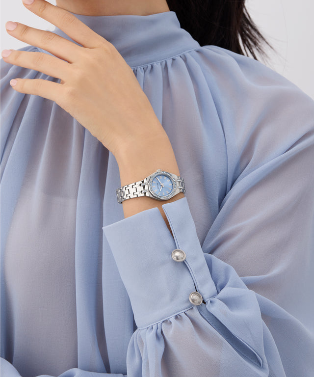 Person wearing a light blue blouse with pearl buttons, holding a silver GUESS Ladies Silver Tone Analog Watch.