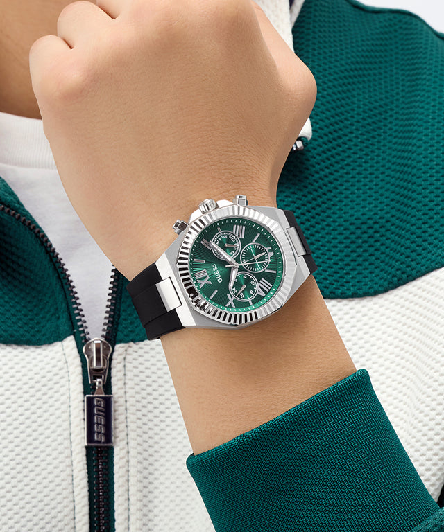 Close-up of a wrist wearing a silver GUESS Mens Equity Black Silver Tone Analog Watch with a green face, against a green and white jacket background.