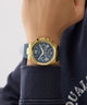 Gold GUESS Mens Equity Blue Gold Tone Analog Watch with blue face on a wrist, wearing a navy sweater.