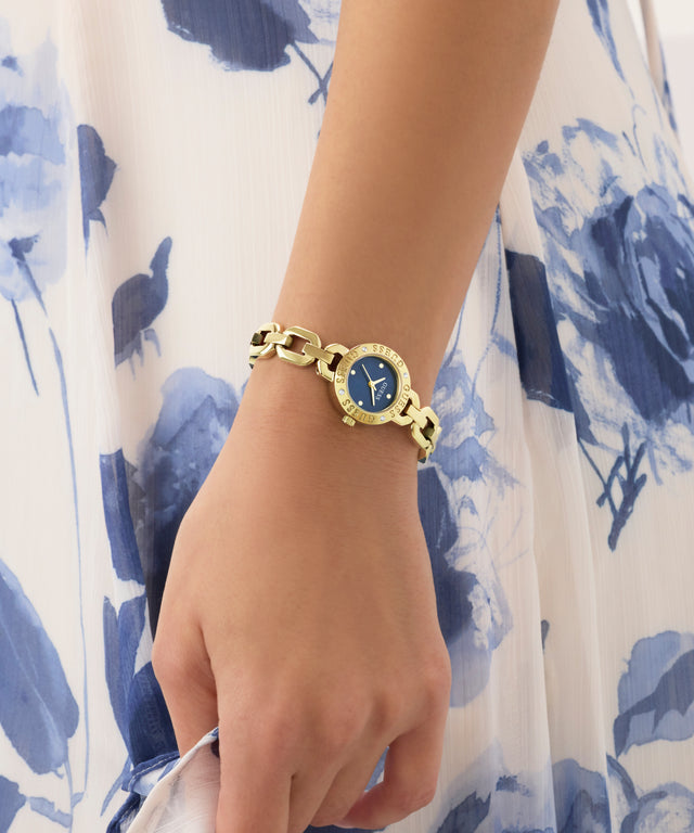 Gold GUESS Ladies Siren Gold Tone Analog Watch with a blue face on a wrist against a floral-patterned background