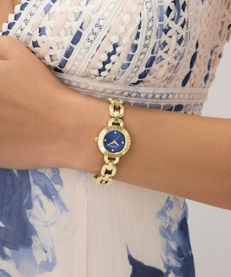 Gold GUESS Ladies Siren Gold Tone Analog Watch with a blue face on a wrist, wearing a white and blue patterned dress.