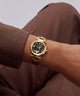 A gold-tone wristwatch with a dark blue dial and gold-tone link bracelet, shown worn on a person's wrist. The person is wearing brown trousers, and the watch brand 'O'NEILL' is visible on the dial.