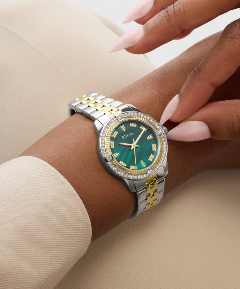 Close-up of a wrist wearing a silver and gold GUESS Ladies Colette 2-Tone Analog Watch with a green face on a beige background.
