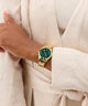 Gold watch with a green face on a wrist against a white textured background