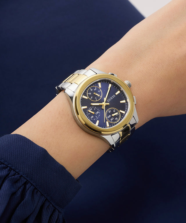Gold and silver GUESS Ladies Birdie 2-Tone Analog Watch on a wrist with a blue background