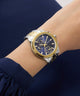 Gold and silver GUESS Ladies Birdie 2-Tone Analog Watch on a wrist with a blue background