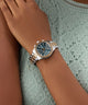 Close-up of a wrist wearing a GUESS Ladies Birdie 2-Tone Analog Watch watch with a detailed face, against a textured green background.