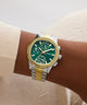 Wrist wearing a Guess watch with a green face and gold accents on a neutral background
