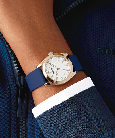Gold GUESS Ladies Blue Rose Gold Tone Analog Watch with blue strap on a wrist, wearing a navy jacket.