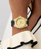 Gold GUESS Ladies White Gold Tone Analog Watch with black strap on a wrist, wearing a white and black outfit.