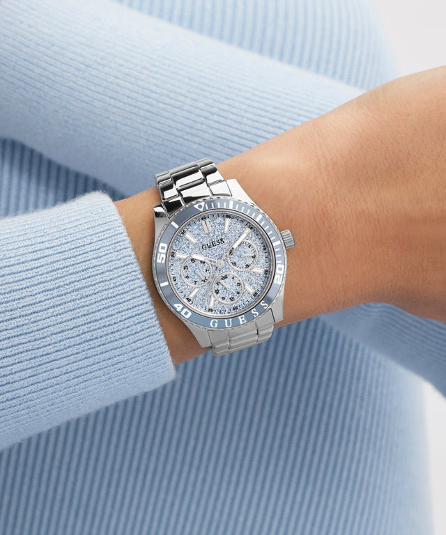 Silver GUESS Ladies Silver Tone Analog Watch with gemstones on a wrist against a light blue background