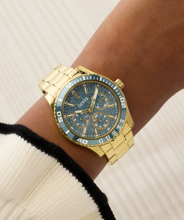 Gold GUESS Ladies Gold Tone Analog Watch with blue face on a wrist against a neutral background