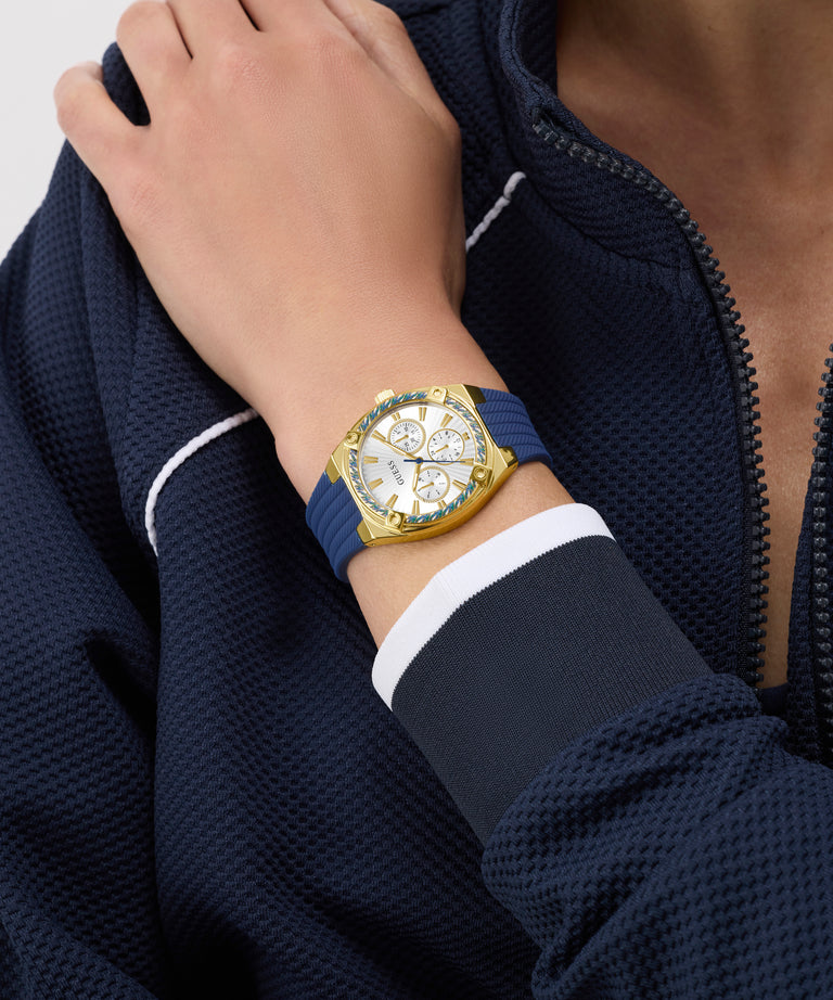 Hand wearing a gold GUESS Ladies Blue Gold Tone Analog Watch with a blue strap on a navy jacket background
