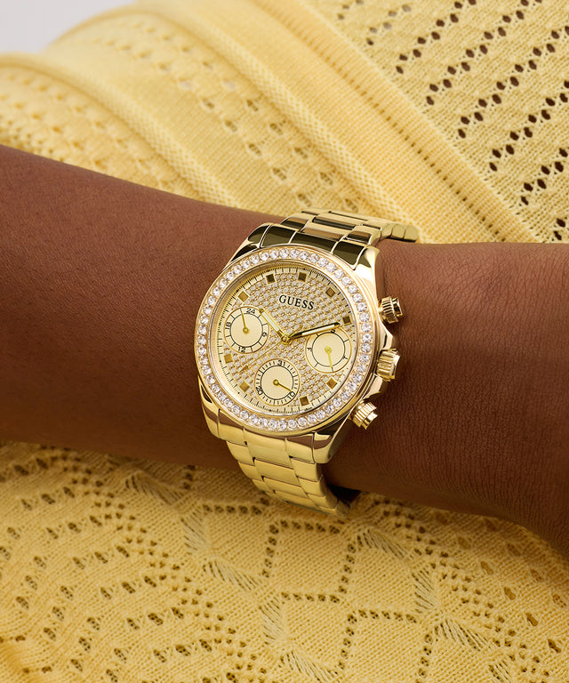 GUESS Ladies Victoria Gold Tone Multi-Function Watch with diamond accents on a wrist against a yellow lace background