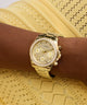 GUESS Ladies Victoria Gold Tone Multi-Function Watch with diamond accents on a wrist against a yellow lace background