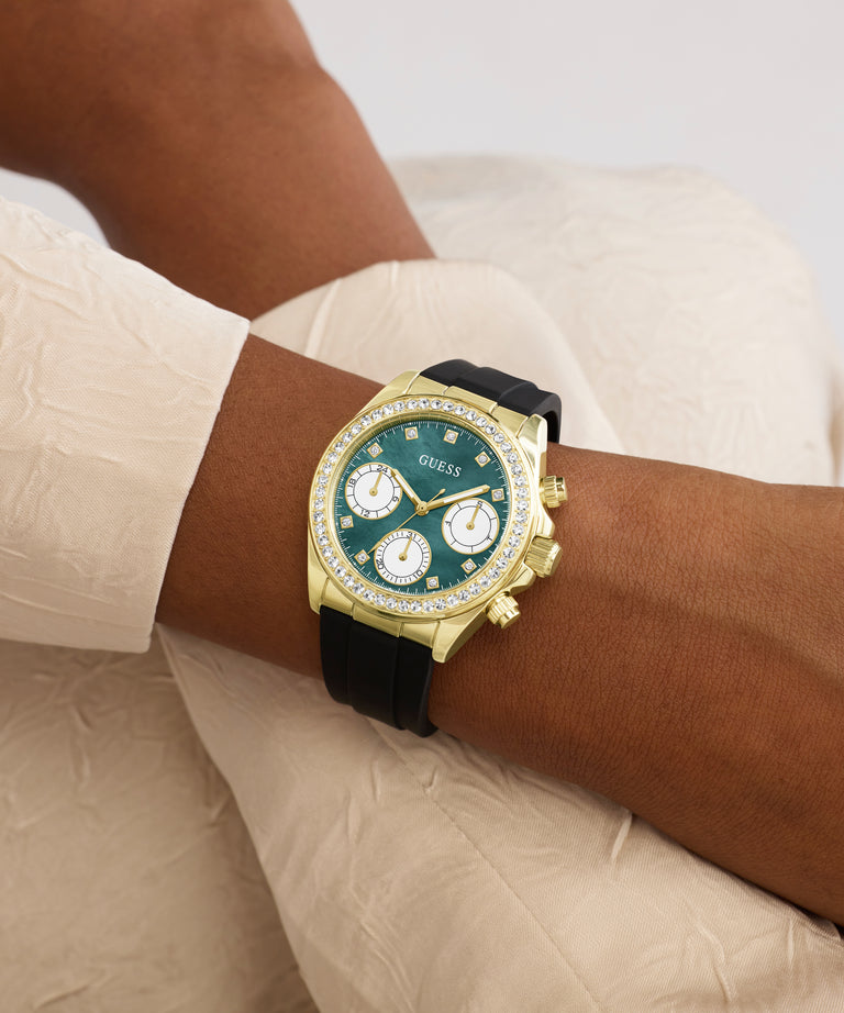 Gold watch with a green face on a wrist, wearing a beige shirt.