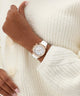 Hand wearing a rose gold watch with a white strap on a white textured background