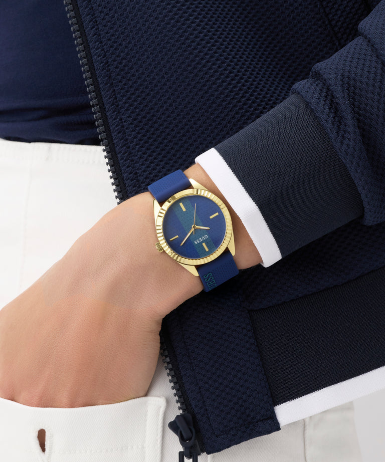 Hand wearing a blue GUESS Ladies Blue Gold Tone Analog Watch with gold accents on a white background