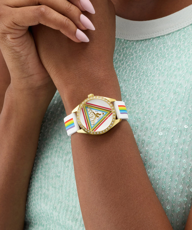 Wrist wearing a GUESS Ladies Pride Limited Edition White Gold Tone Analog Watch with a colorful band and decorative face, against a light green textured background.