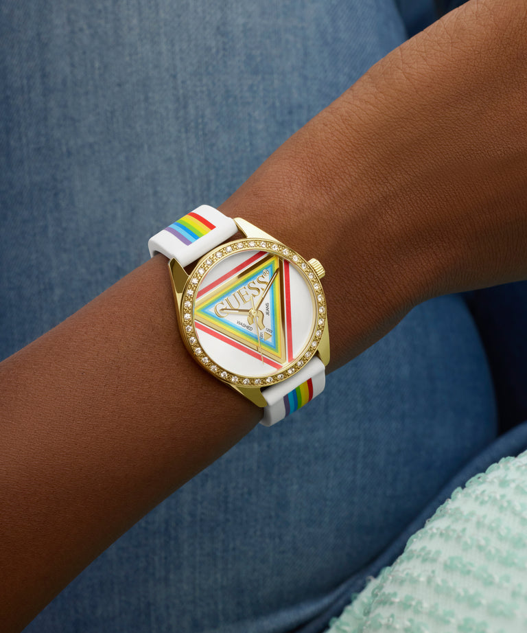 Wrist wearing a GUESS Ladies Pride Limited Edition White Gold Tone Analog Watch with a colorful face and rainbow strap against a blue background
