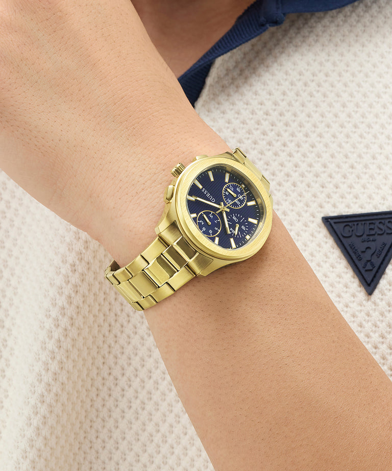 Gold GUESS Mens Palmer Gold Tone Analog Watch with blue face on a wrist, brand logo in the background