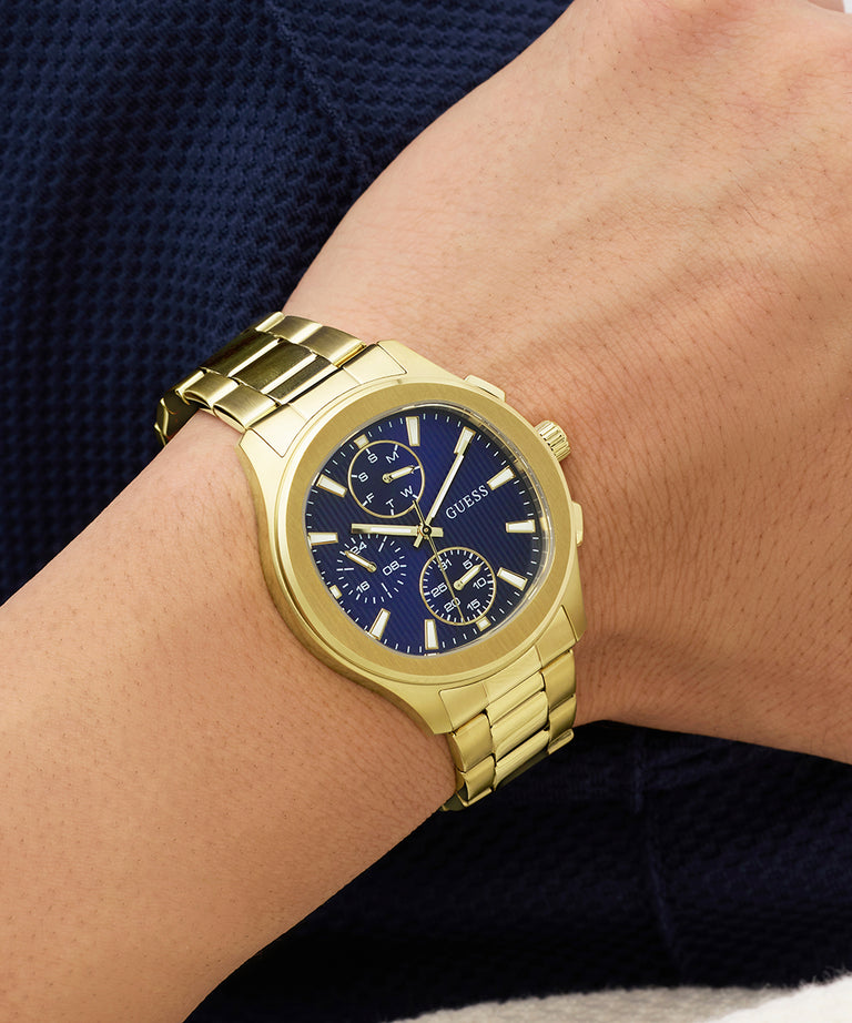 Gold GUESS Mens Palmer Gold Tone Analog Watch with a blue face on a wrist against a dark background