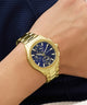Gold GUESS Mens Palmer Gold Tone Analog Watch with a blue face on a wrist against a dark background