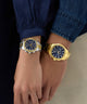 Two wrists wearing gold watches with blue faces against a dark background