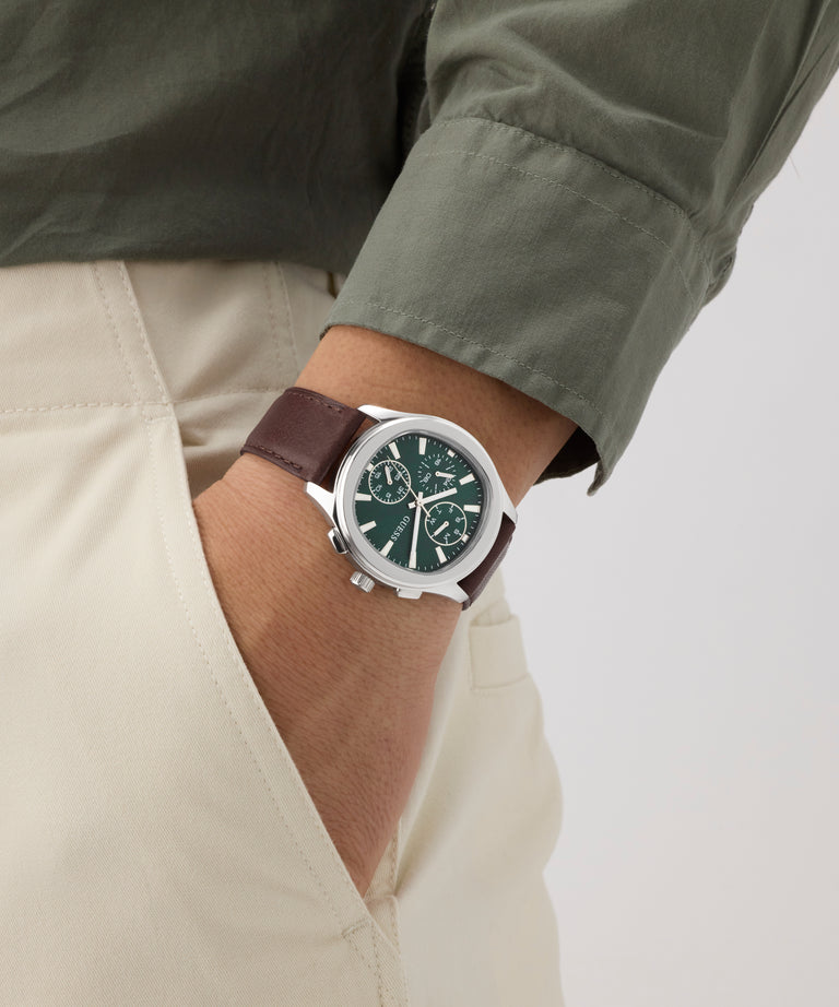 Person wearing a GUESS Mens Palmer Light Brown Silver Tone Analog Watch with a green face and brown strap on a neutral background