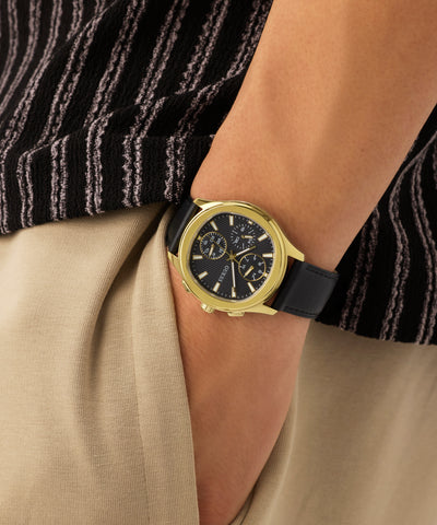 Gold GUESS Mens Palmer Black Gold Tone Analog Watch with black strap on a wrist, wearing beige pants and a black and white striped shirt.