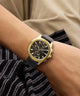 Gold watch with black strap on a wrist, wearing a striped shirt.