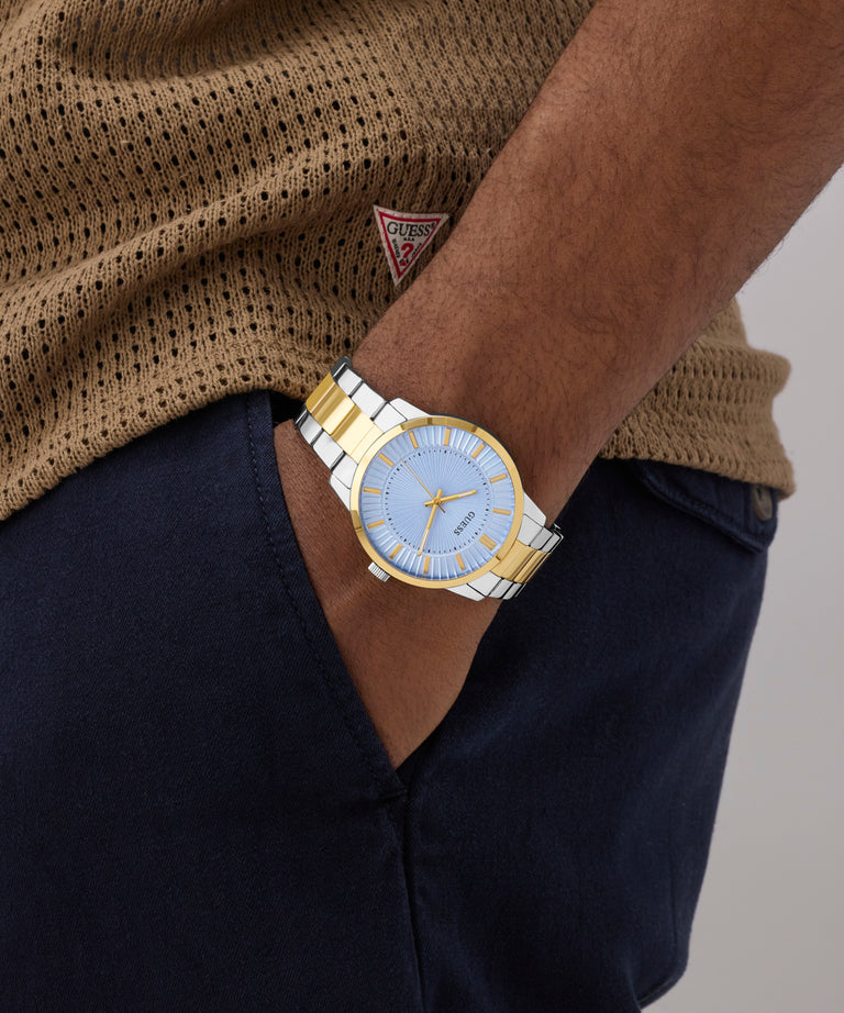 Person wearing a gold and silver watch with a blue face, brown sweater, and navy pants.