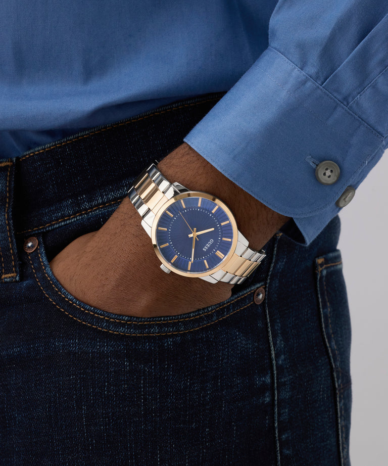 Person wearing a blue shirt and dark jeans with a gold GUESS Mens 2-Tone Analog Watch on a gray background