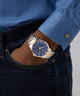 Person wearing a blue shirt and dark jeans with a gold GUESS Mens 2-Tone Analog Watch on a gray background