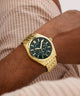 Gold GUESS Mens Gold Tone Analog Watch with a dark green face on a wrist, against a neutral background