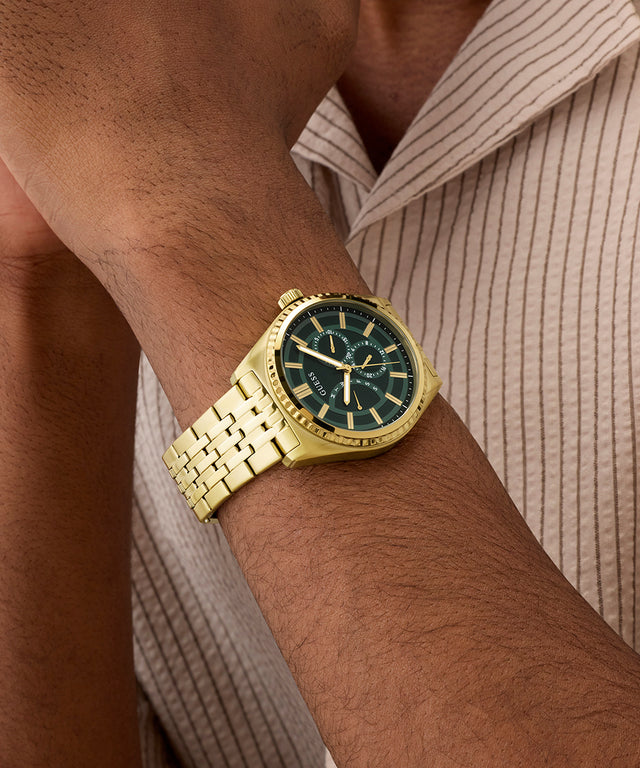 Gold GUESS Mens Gold Tone Analog Watch with a green face on a person's wrist, wearing a light brown shirt.
