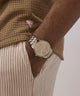Person wearing a GUESS Mens Coffee Analog Watch with a brown sweater and beige pants on a neutral background