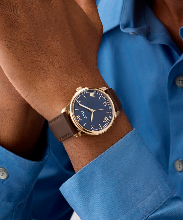 Close-up of a wrist wearing a GUESS Mens Dark Brown Rose Gold Tone Analog Watch with a blue face and brown strap, against a blue shirt background.