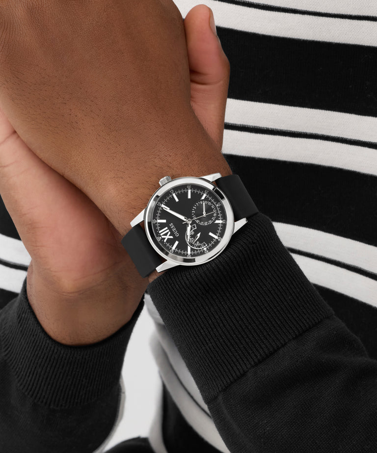Hand wearing a black GUESS Mens Black Silver Tone Analog Watch with a detailed face, against a blurred background.