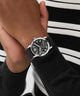Hand wearing a black GUESS Mens Black Silver Tone Analog Watch with a detailed face, against a blurred background.