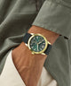 Person wearing a watch with a gold case and green dial on a wrist, with a blurred background.