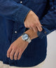 Person wearing a silver GUESS Mens Arthur Silver Tone Analog Watch with a blue face on a plain background