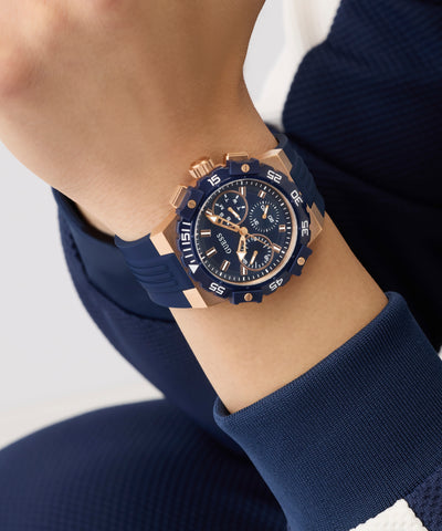 Person wearing a blue GUESS Mens Midnight Blue Rose Gold Tone Analog Watch with rose gold accents on a white background