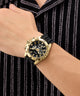 Gold wristwatch with black strap on a person's wrist against a black and gray striped background