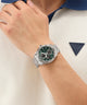 Person wearing a silver watch with a green face on a white background, featuring a Guess logo.