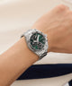 Silver watch with a green face on a wrist against a neutral background