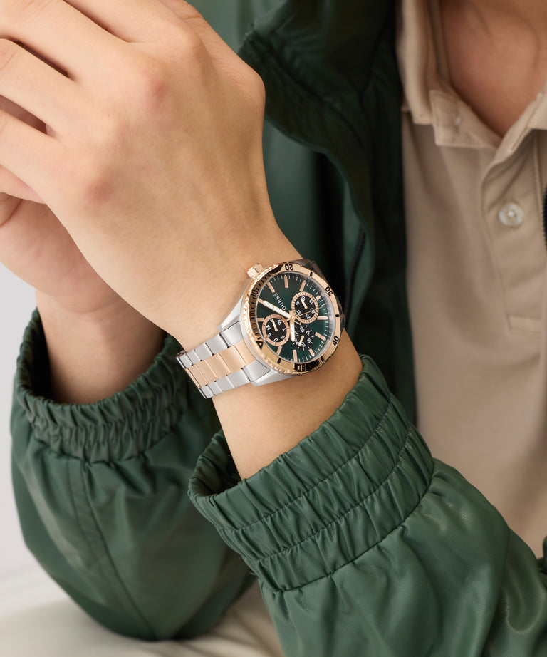 Close-up of a wrist wearing a silver and rose gold watch with a green jacket sleeve.