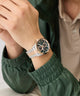 Close-up of a wrist wearing a silver and rose gold watch with a green jacket sleeve.